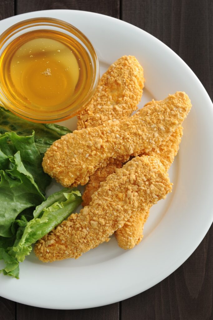 Giada Crunchy Parmesan Chicken Tenders with a side of honey dip and lettuce on a white plate.