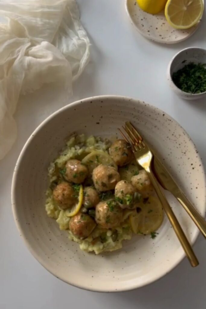 Giada De Laurentiis Chicken Piccata Meatballs served with lemon slices and parsley in a white bowl.