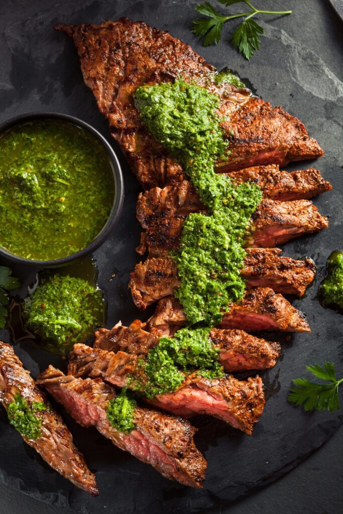 Sliced grilled steak with Giada De Laurentiis chimichurri sauce on a slate platter.