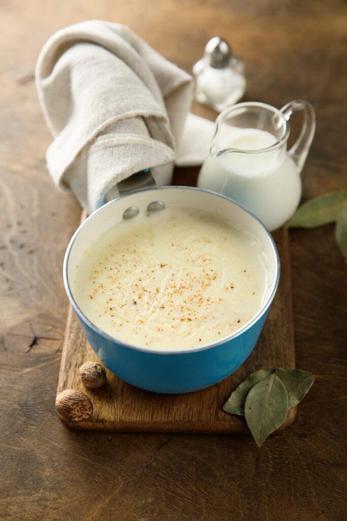 Pot of creamy bechamel sauce with milk and nutmeg on a wooden board.