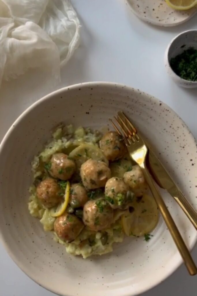 Chicken Piccata Meatballs in a white bowl with lemon slices and a gold fork and knife.