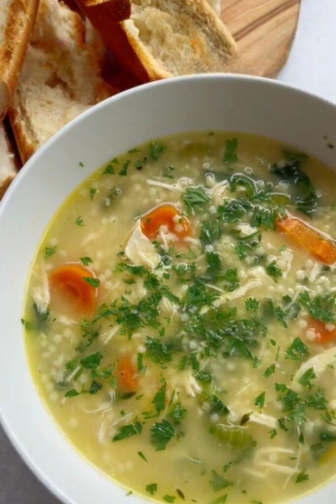 hicken Pastina soup in a white bowl with bread slices on a wooden board.