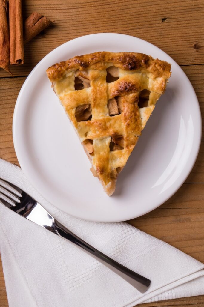 Overhead view of a slice of apple Parmesan pie with a lattice crust on a white plate, next to a fork and napkin.