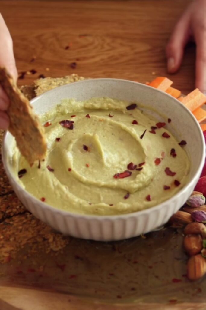 A cracker dipping into a bowl of avocado and white bean dip, surrounded by carrot sticks.