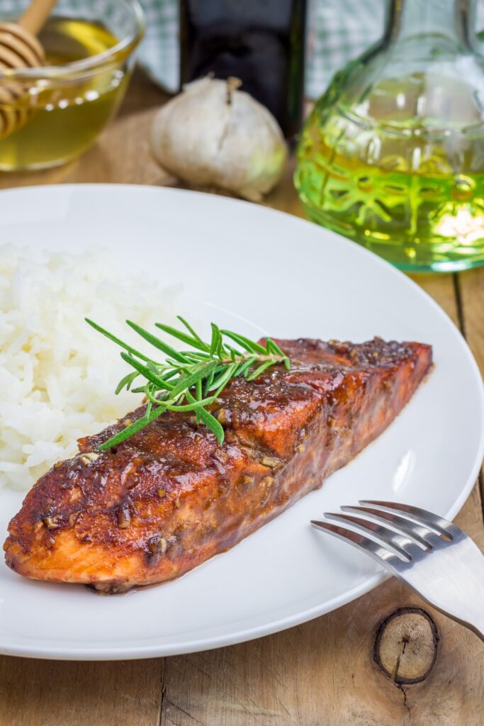 Giada Balsamic-glazed Salmon fillets in a baking tray garnished with rosemary, surrounded by ingredients like olive oil and garlic.
