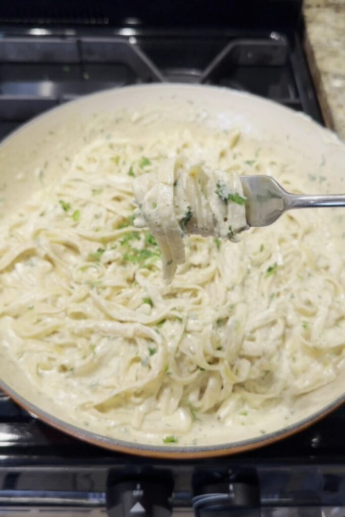 A fork holding a bite of Giada De Laurentiis Fettuccine Alfredo above a pan of pasta.