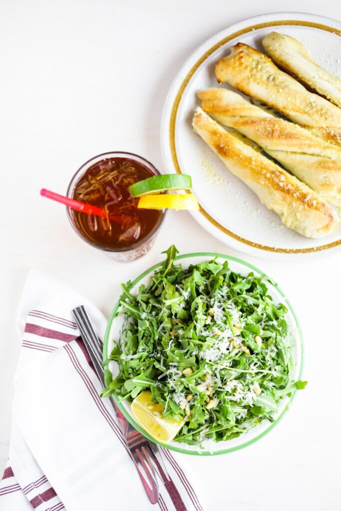 Giada Candied Lemon and Arugula Salad with pine nuts, grated cheese, and a lemon wedge, served with a drink and breadsticks.