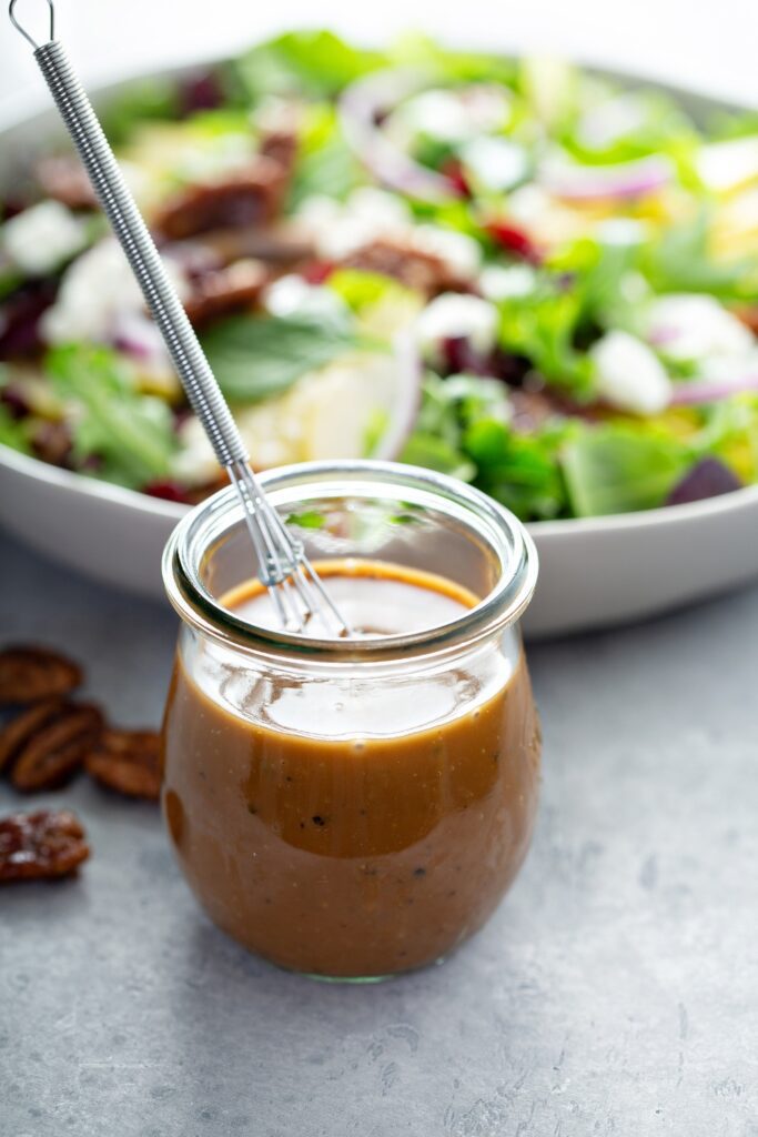 Jar of balsamic vinaigrette with a whisk, salad blurred in the background.