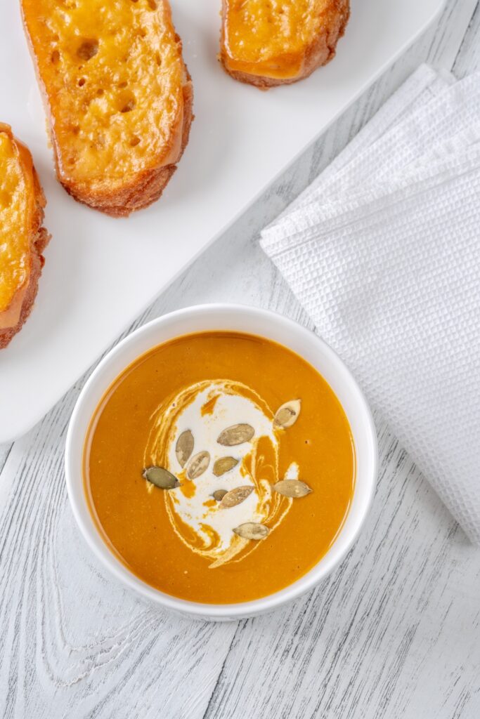 A bowl of Giada Butternut Squash Soup garnished with cream and pumpkin seeds, served with Fontina cheese crostini.