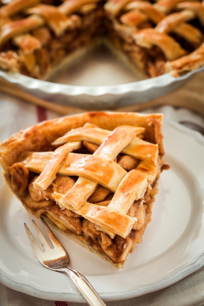 A slice of apple Parmesan pie with a lattice crust on a white plate, with the rest of the pie in the background.