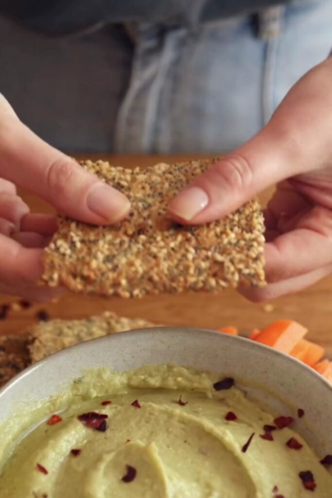 Hands breaking a seed-covered cracker with a bowl of avocado and white bean dip in the background.