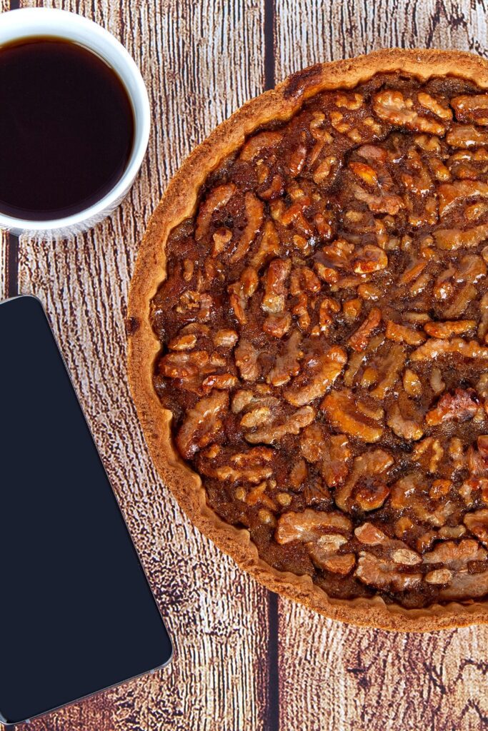 Brownie walnut pie next to a cup of coffee and a smartphone on a wooden table.