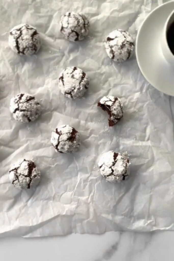 Multiple Giada De Laurentiis chocolate amaretti cookies dusted with powdered sugar on parchment paper with a cup of coffee.