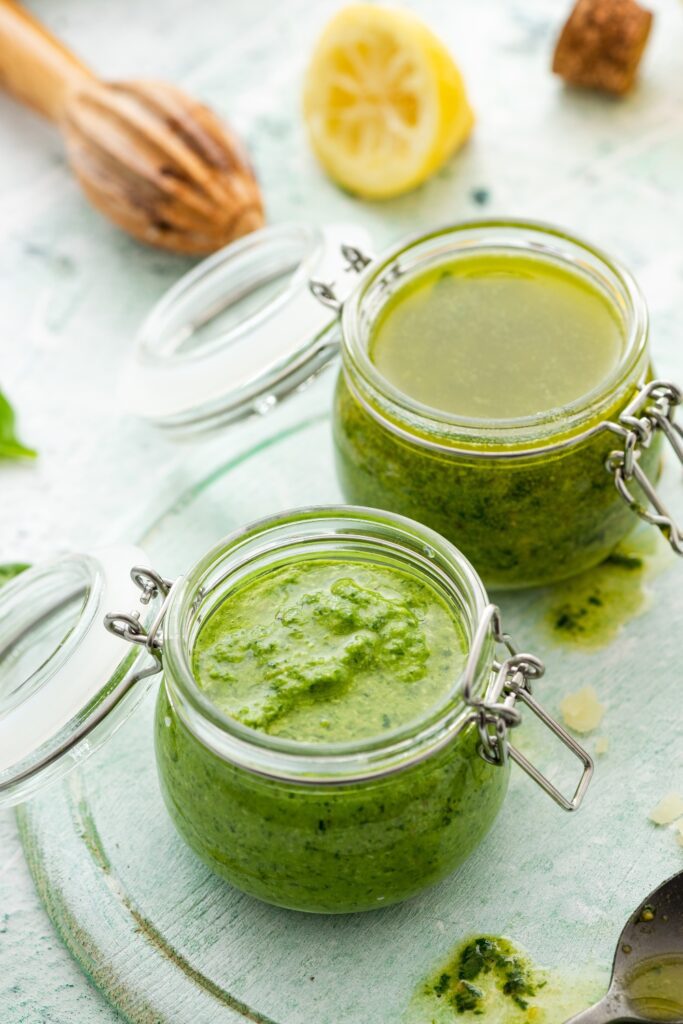 Two glass jars of basil pesto on a light green surface with a lemon half and a wooden juicer in the background.