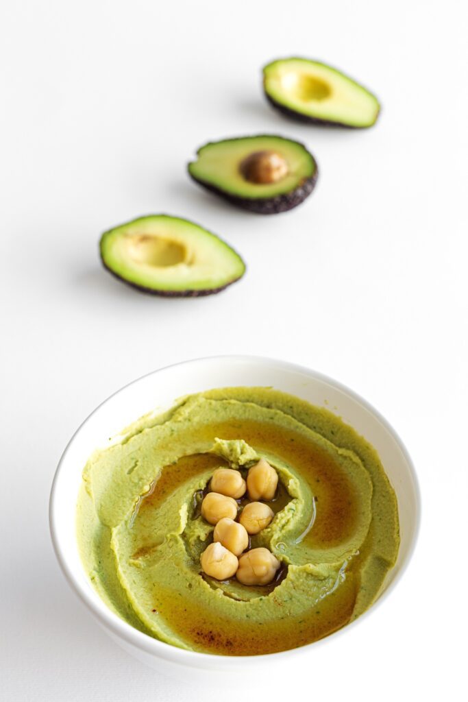 A bowl of Giada Avocado Hummus with chickpeas on top, with avocado halves in the background.