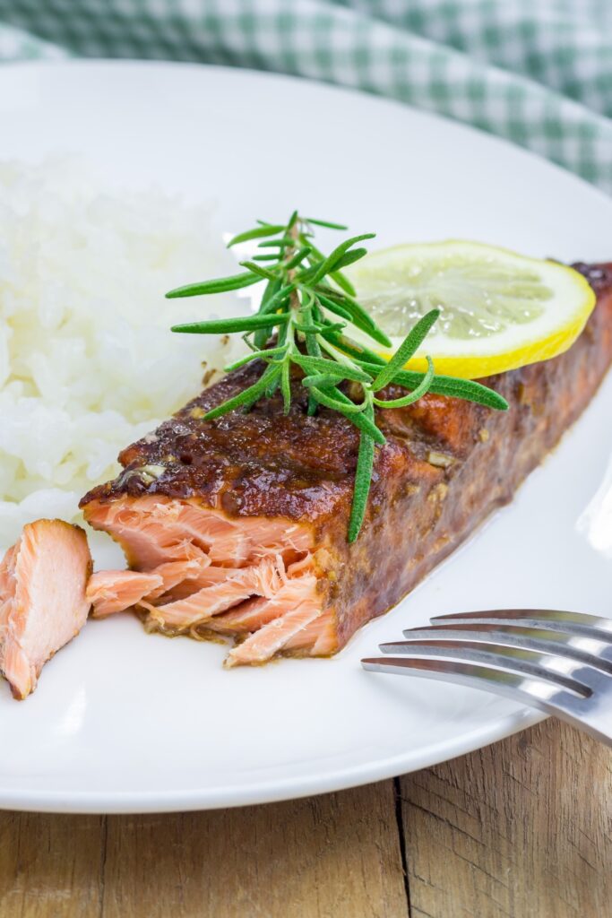 A piece of Giada Balsamic-glazed Salmon with rice, a sprig of rosemary, and a garlic bulb in the background.